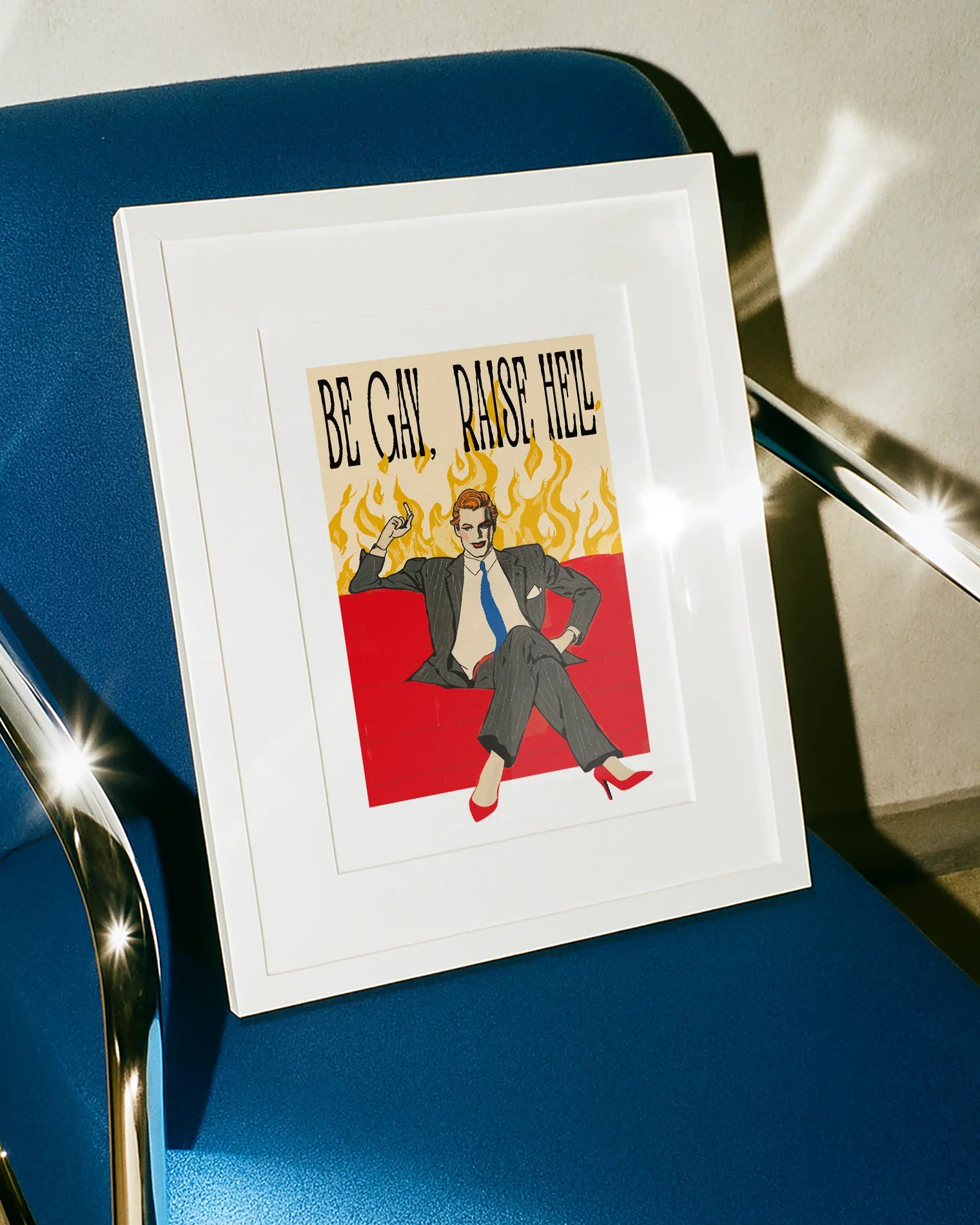 A framed poster featuring a man in make up, red high heels, sitting on a couch smoking, surrounded by massive flames, with the text "BE GAY, RAISE HELL".