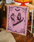 A poster featuring a female devil sitting on the edge of a coffin, while a female vampire sits inside. They hold each other’s hands romantically, creating a mysterious and intimate atmosphere.