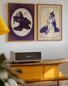 A poster with a witch tenderly holding her lover, surrounded by a spiderweb-infused frame and a large white full moon with flying bats in the background.