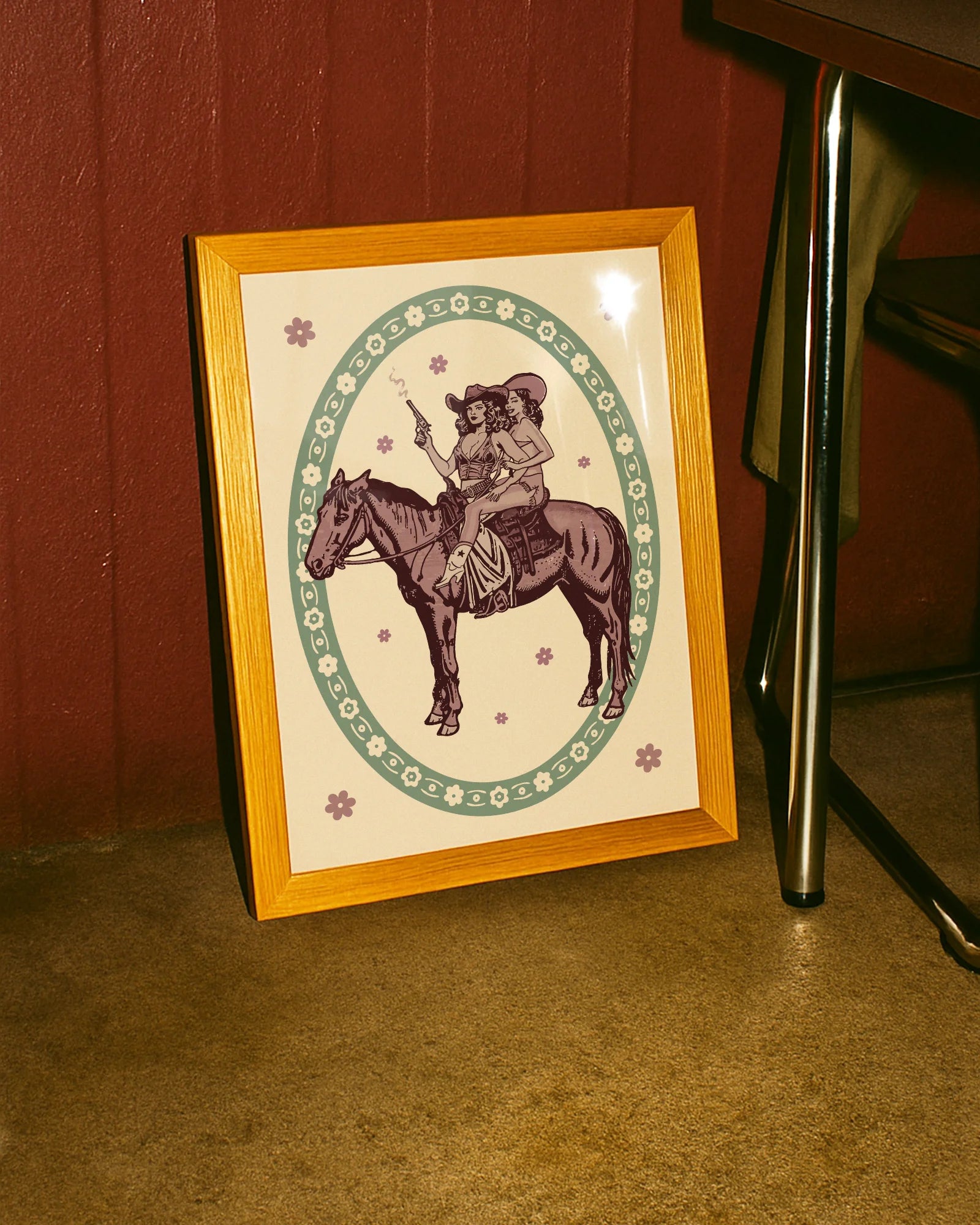A framed poster featuring two lesbian cowgirls sitting on a horse, surrounded by a retro-style floral frame.