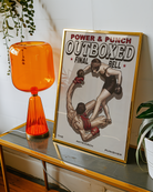 A framed poster featuring a female boxer completely dominating the ring, knocking down her male opponent, with the texts: “POWER & PUNCH”, “OUTBOXED”, “FINAL”, and “THE PATRIARCH PUNISHER.”